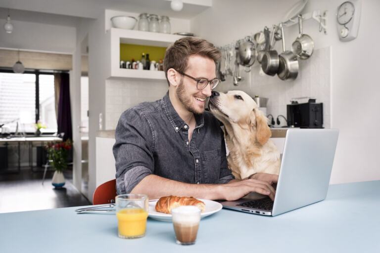 Mann am Laptop am Küchentisch, ein Golden Retriever legt den Kopf auf seine Schulter